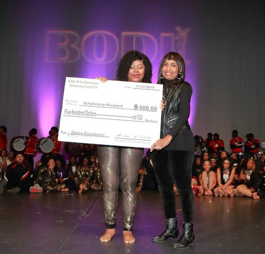 Two people stand on stage holding a large $500 scholarship check. The background features "BODI" in illuminated letters and a group of performers seated. The event date is 11/17/2019.